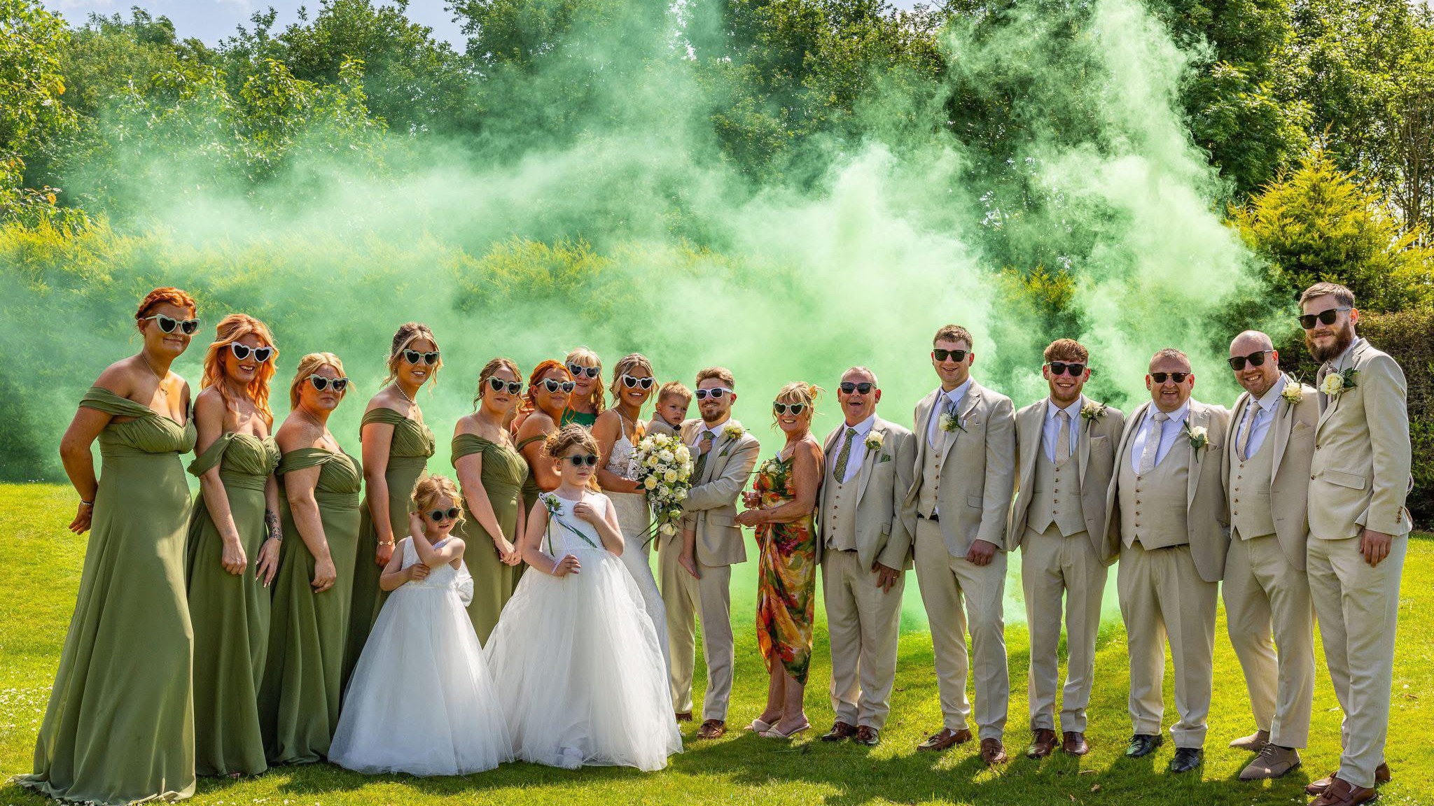 A bride and groom holding their young son are flanked by their bridesmaids and groomsmen on The Lawn of  The Manor; green smoke billows behind them.