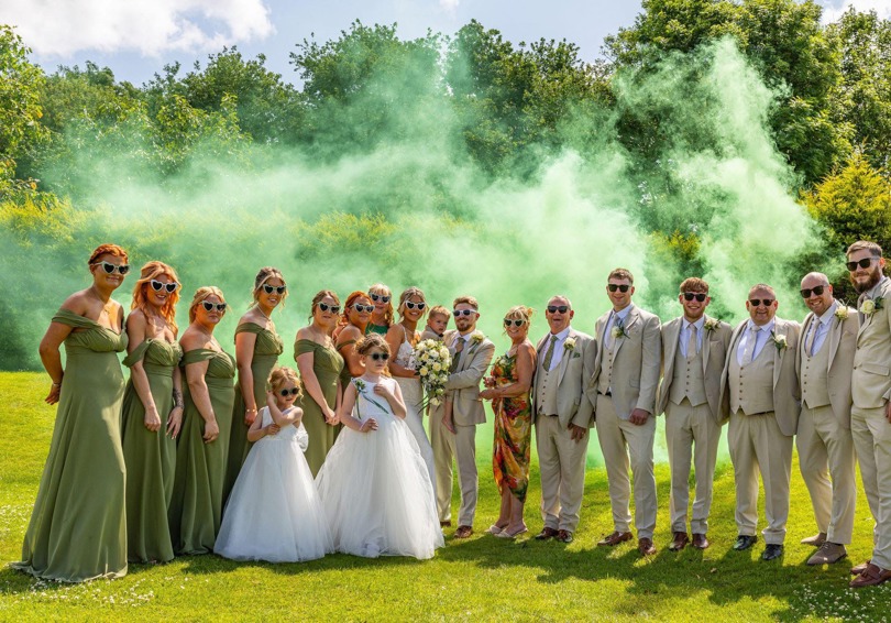 A bride and groom holding their young son are flanked by their bridesmaids and groomsmen on The Lawn of  The Manor; green smoke billows behind them.