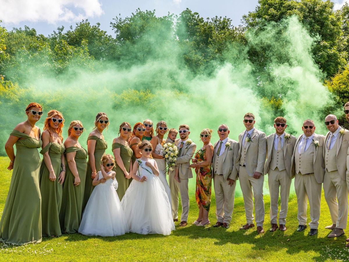 A bride and groom holding their young son are flanked by their bridesmaids and groomsmen on The Lawn of  The Manor; green smoke billows behind them.