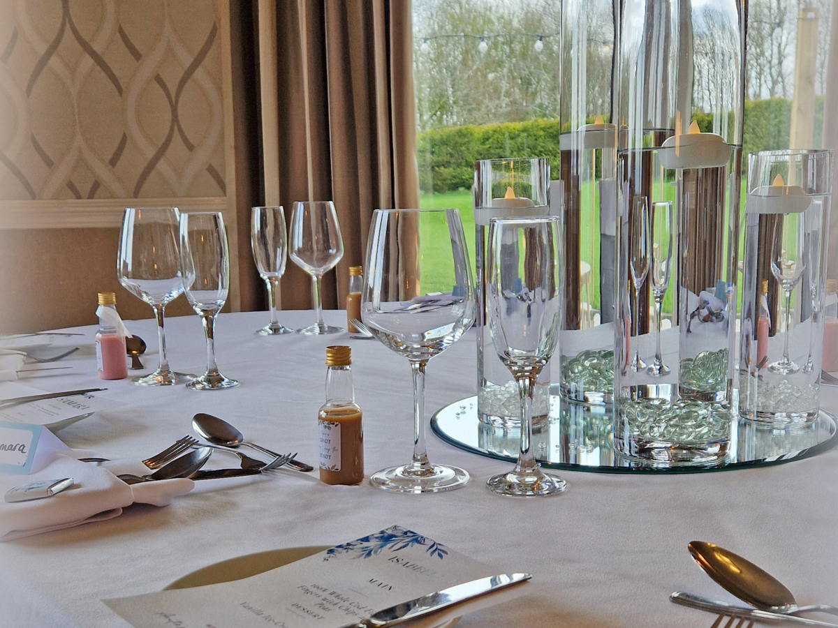 A dining table at The Manor set for a wedding breakfast, the centrepiece consists of tall glass vases, the view over The Lawn can be seen in the background.