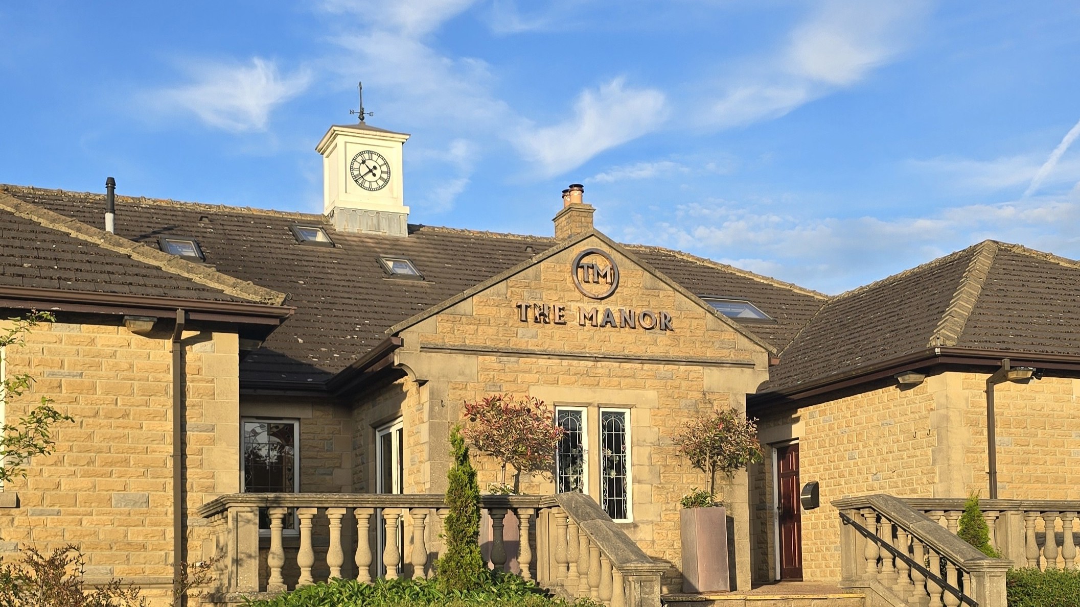 The main entrance of The Manor, showing the front steps, stone balustrade, the clocktower and the gold logo.