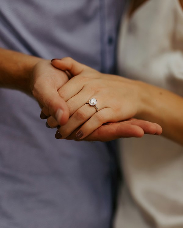 A female left hand placed on a male right hand, there is a diamond engagement ring on her third finger.