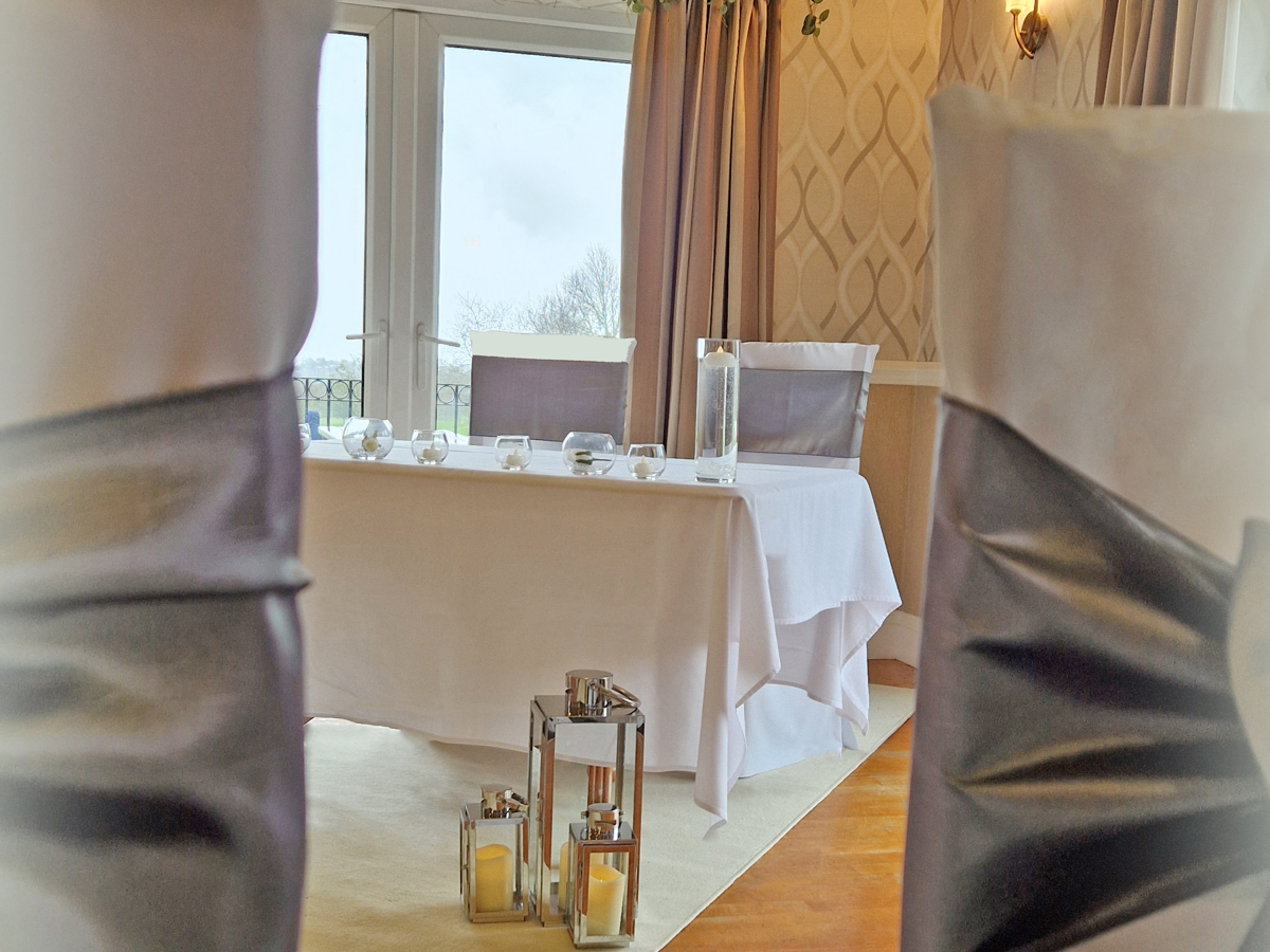 A view of the table at the end of the aisle in the High Garden room at The Manor taken between two chairs. Silver lanterns sit in front of the table.