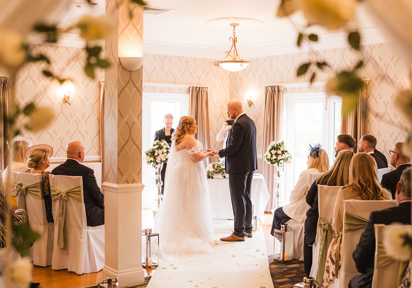 A wedding ceremony taking place in High Garden at The Manor. The view is through a floral archway, the bride and groom face each other at the end of the aisle.
