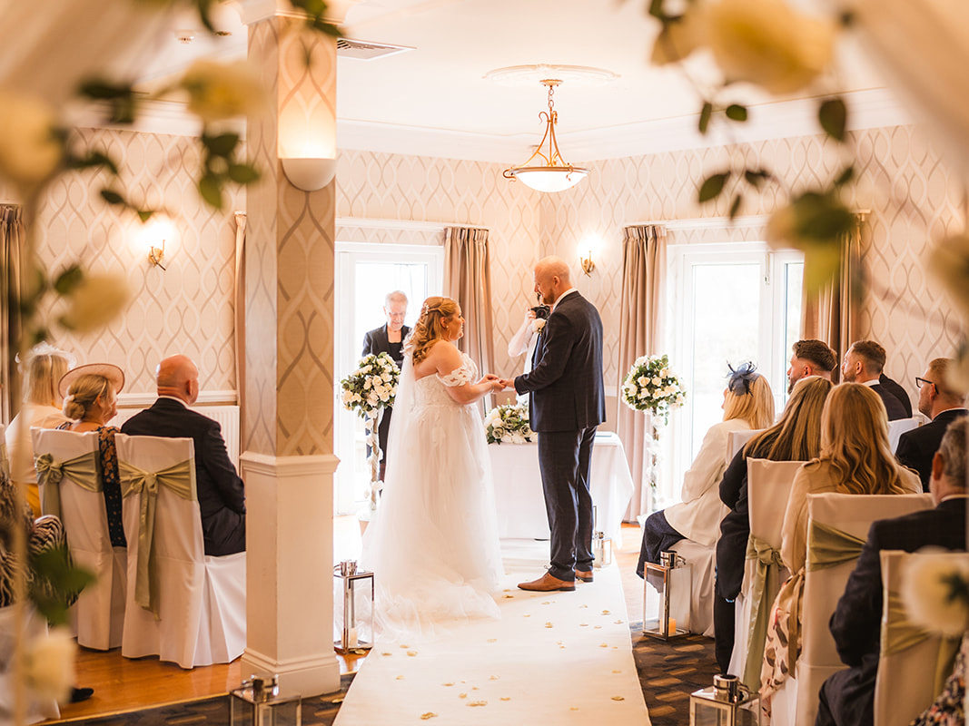 A wedding ceremony taking place in High Garden at The Manor. The view is through a floral archway, the bride and groom face each other at the end of the aisle.