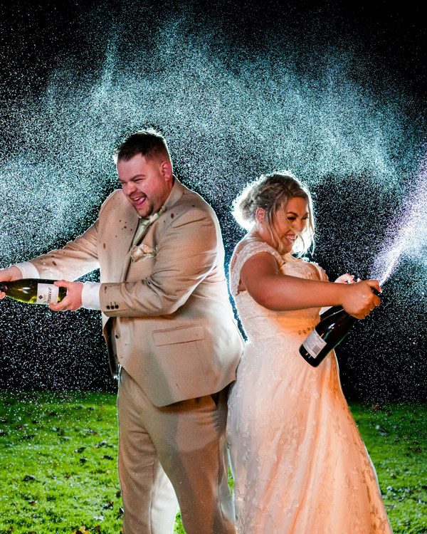 A night shot of a bride and groom spraying prosecco on The Lawn of The Manor.