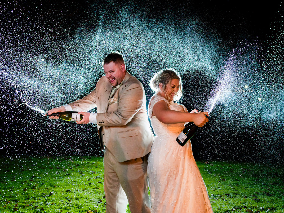 A night shot of a bride and groom spraying prosecco on The Lawn of The Manor.