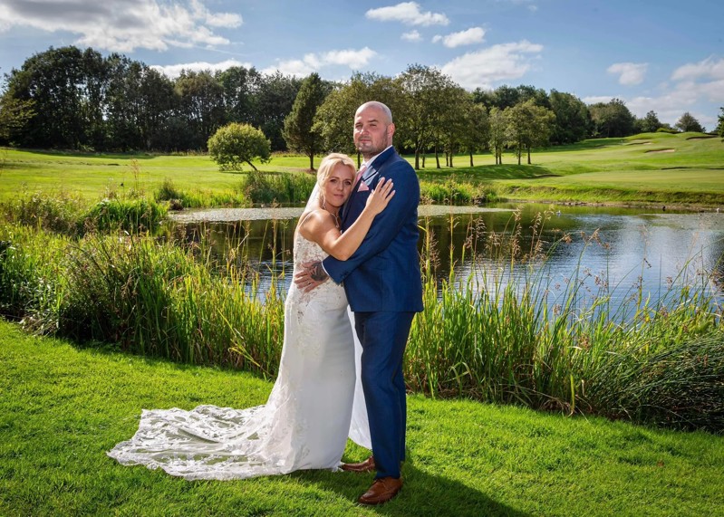 A bride and groom embrace in front of The Lake at The Manor Leeds on a sunny day.