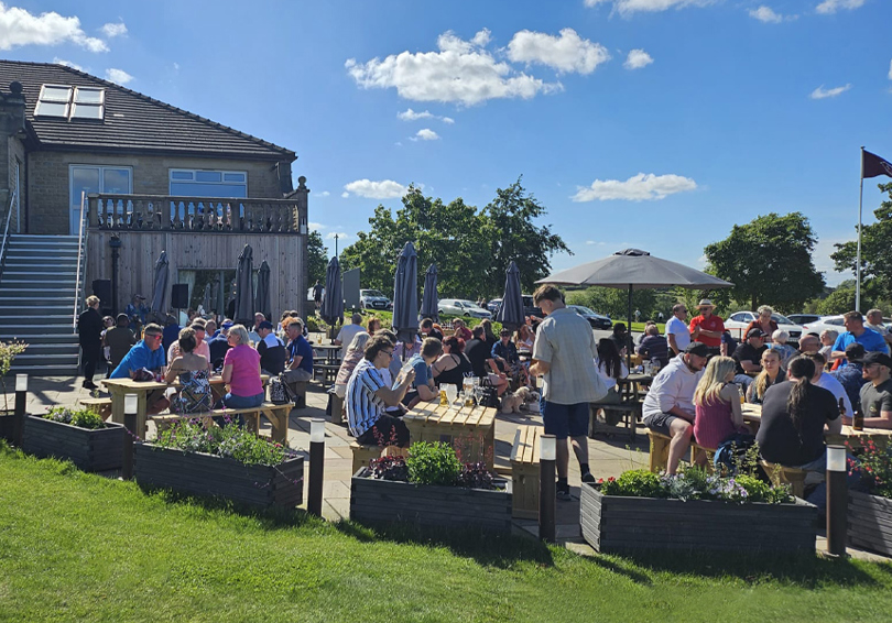 The patio area at Cook House Bar & Kitchen Leeds on a sunny day. The sky is bright blue with a few isolated clouds. The wooden picnic-style tables are occupied by a variety of people. In the foreground there are wooden planters.