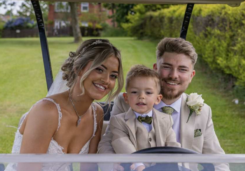 A bride and groom smiling from a golf buggy, their young son is sitting on his father's lap.