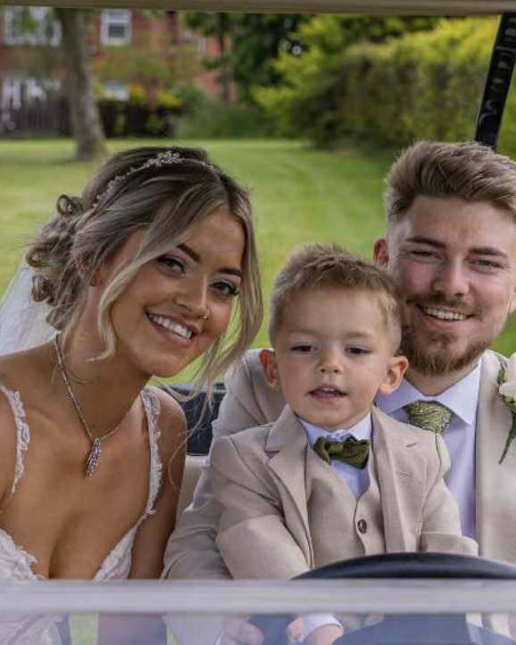 A bride and groom smiling from a golf buggy, their young son is sitting on his father's lap.