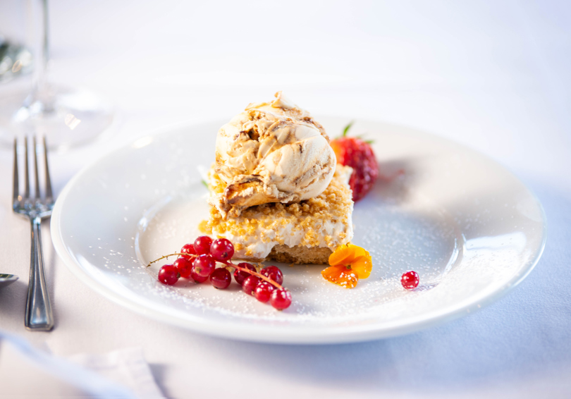 Cheesecake on a plate, topped with ice cream, red currents and an edible flower sit beside.