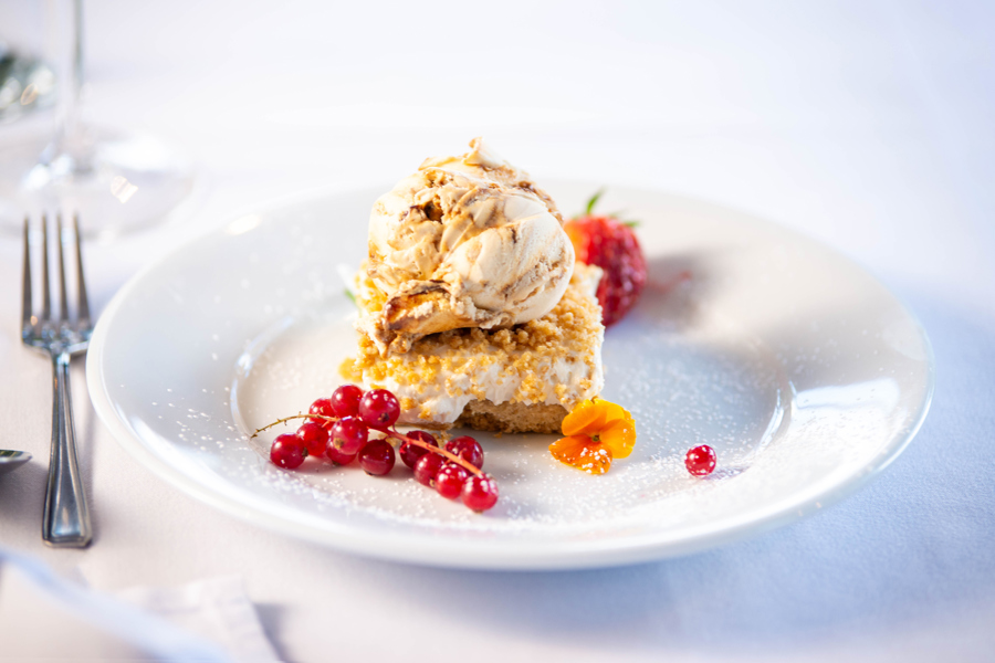 Cheesecake on a plate, topped with ice cream, red currents and an edible flower sit beside.