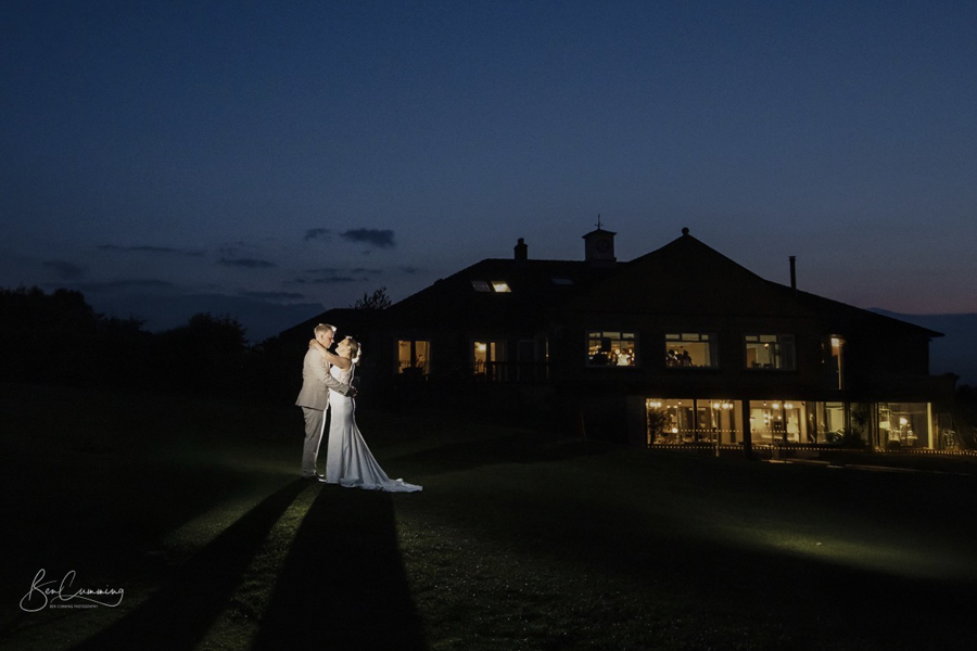 A night shot of bride and groom embracing with the rear aspect of The Manor main building in the background.