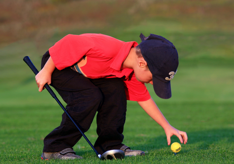 A young boy placing a yellow golf ball on a tee with his left hand, he has a golf club in his right hand.