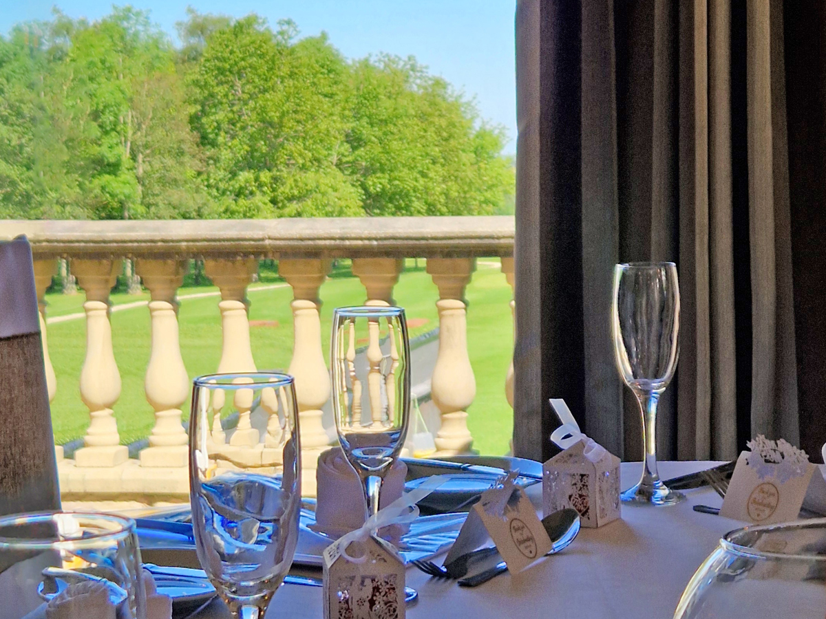 A view over a table set for a wedding breakfast in the High Garden room at The Manor, looking out of the open doors towards the trees beyond the stone balustrade.