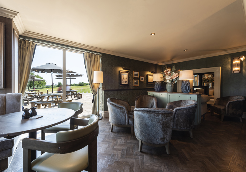 A photo of the snug area of Cook House Bar & Kitchen. A dining table and chairs are on the left of the image and a group of four armchairs are towards the centre in front of a console table holding a large vase of flowers and two lamps. A double sided stove can be seen beyond the table. Picnic benches with parasols can be seen through the large full length windows.