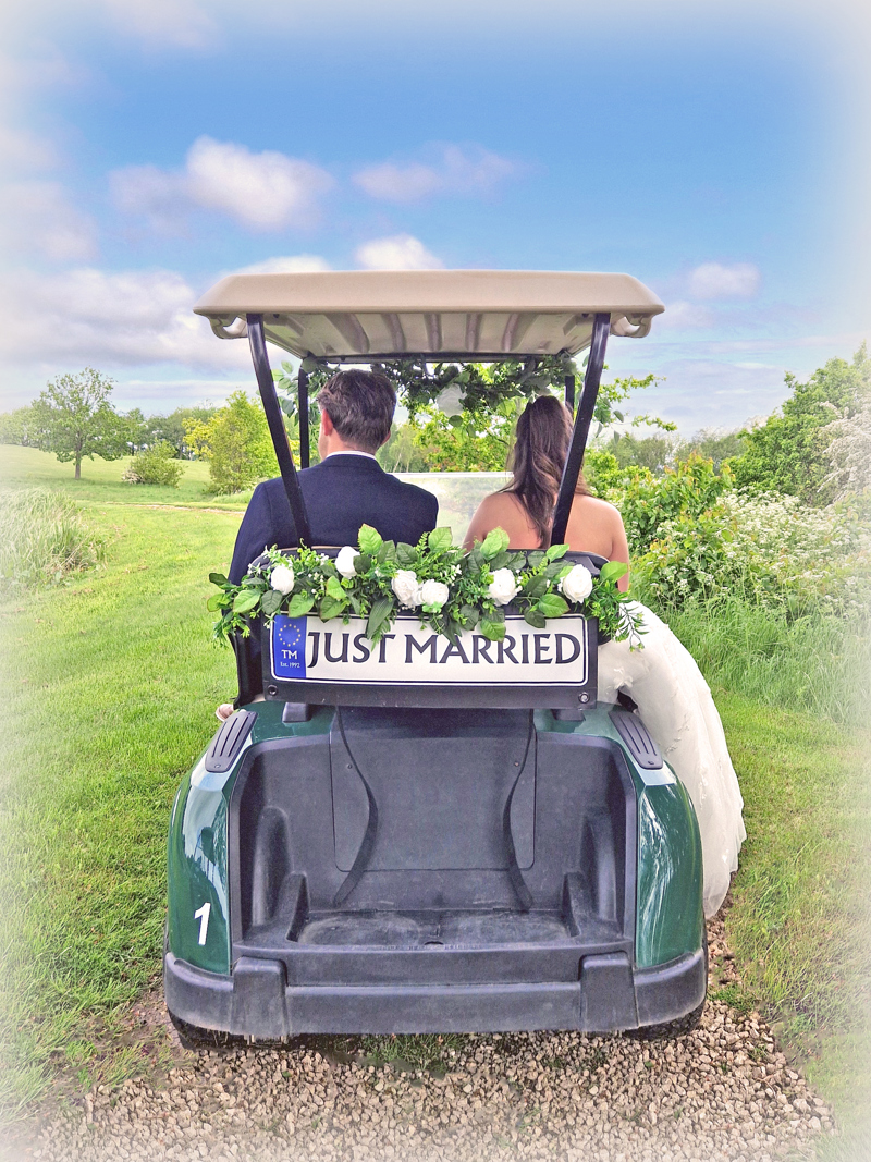 A rear view of a bride and groom sitting in a golf buggy decorated with a "Just Married" sign and white flowers.