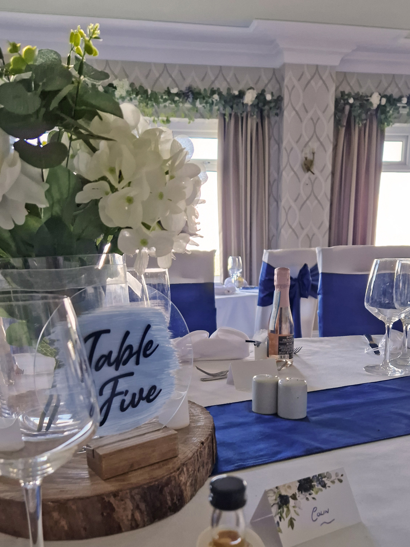 A dining table at The Manor set for a wedding breakfast, the table runner and chair sashes are royal blue, white hydrangeas sit in a vase on a wood slice in front of a sign saying "Table Five".