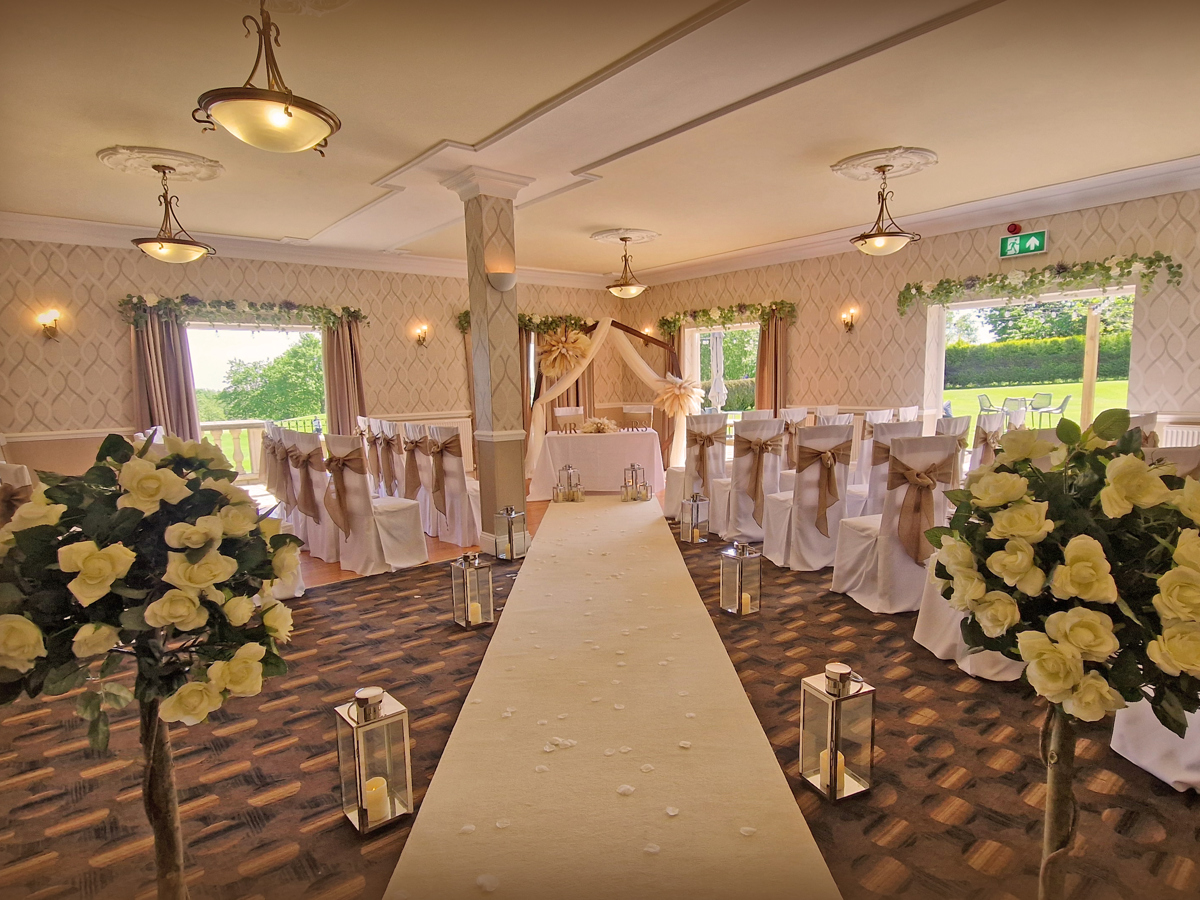 The High Garden room at The Manor set up for a wedding ceremony with an aisle runner, silver lanterns, hessian chair sashes and white rose trees.