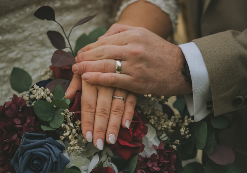 The left hands of a male and female, both wearing wedding rings, placed on her burgundy bouquet.