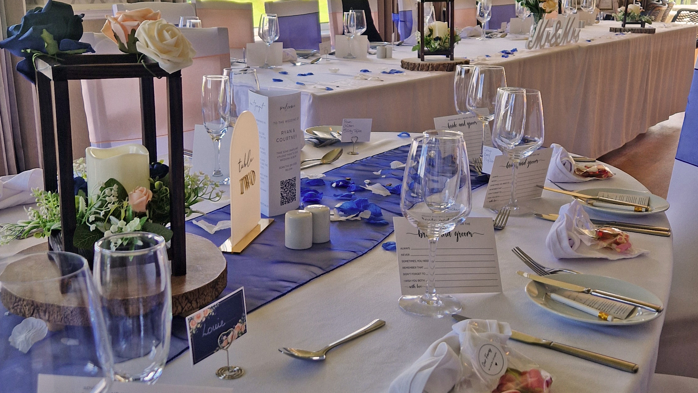 A dining table at The Manor set for a wedding breakfast, the white linen napkins are folded into hearts and the table runner is dark blue. The long top table can be seen in the background and grass and trees can be seen through the windows.