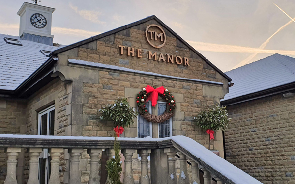The main entrance of The Manor, showing the stone balustrade, the clocktower and the gold logo, a large Christmas wreath with a red bow hangs beneath the sign.
