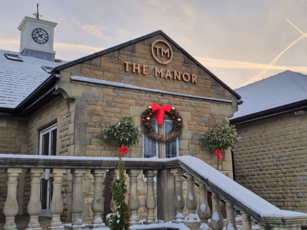 The main entrance of The Manor, showing the stone balustrade, the clocktower and the gold logo, a large Christmas wreath with a red bow hangs beneath the sign.