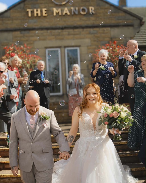 A bride and groom holding hands outside The Manor's main entrance, their guests are blowing bubbles over them.