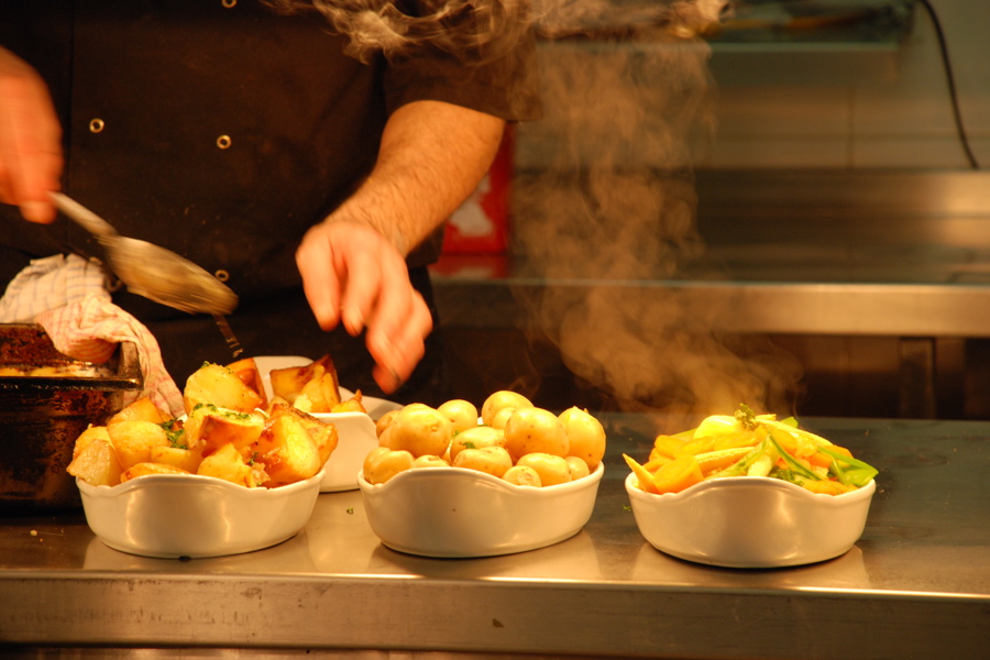 Steaming vegetables, new potatoes and roast potatoes in dishes with a chef in the background.