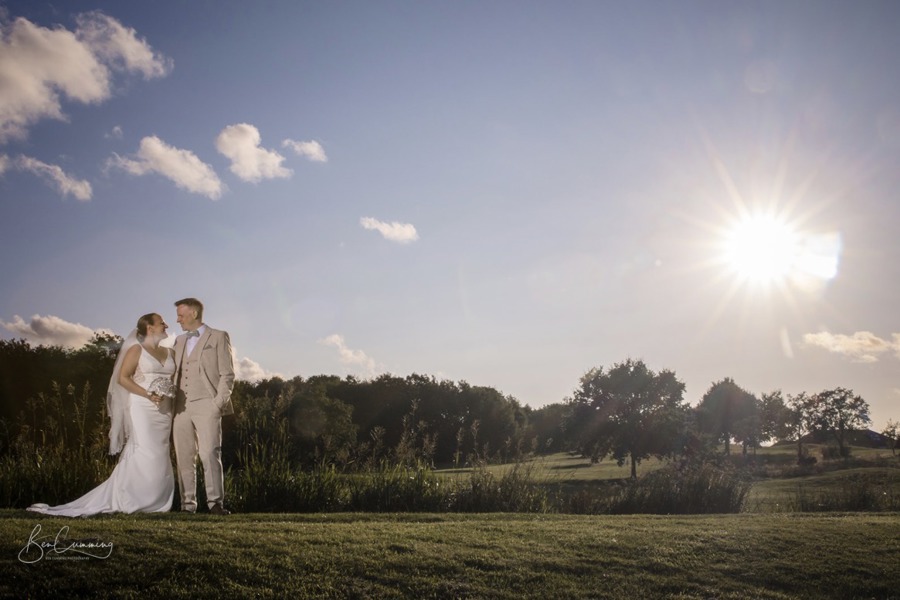 A bride and groom gaze at each other, standing beside The Lake at The Manor Leeds, the sun shines dramatically to the top right.