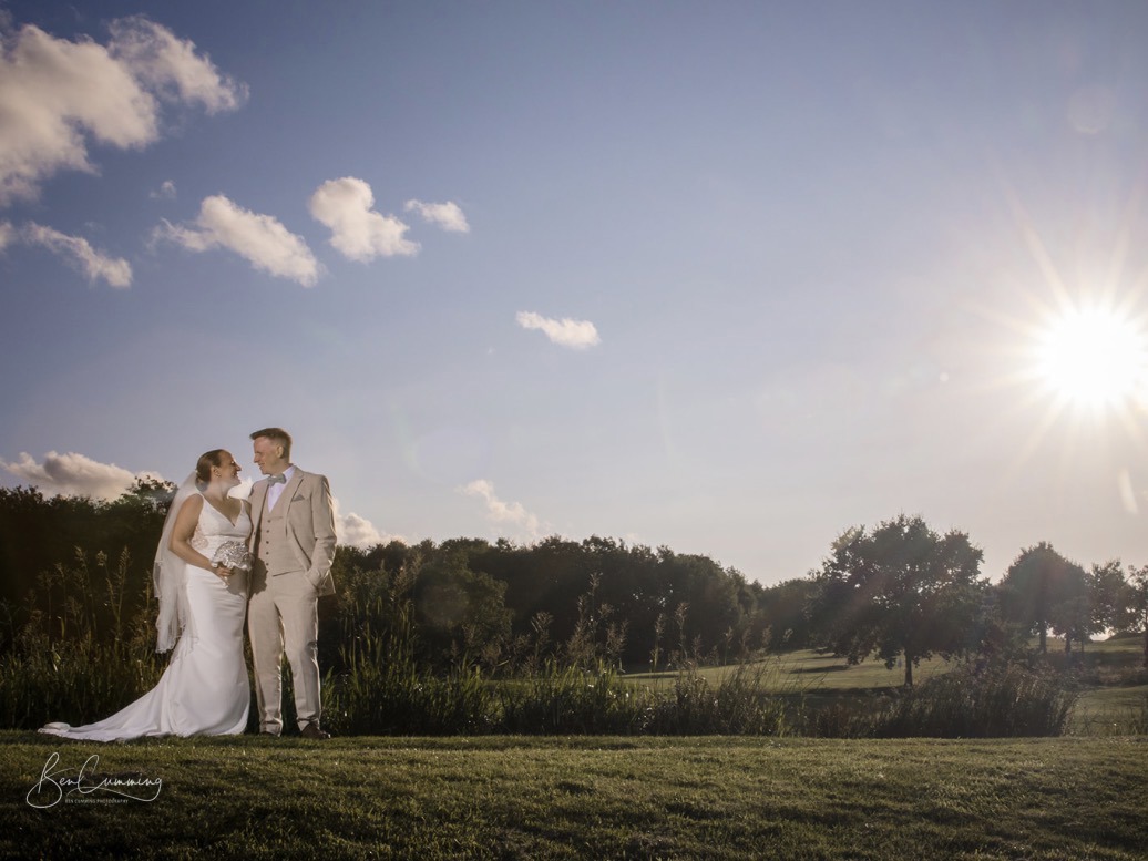 A bride and groom gaze at each other, standing beside The Lake at The Manor Leeds, the sun shines dramatically to the top right.