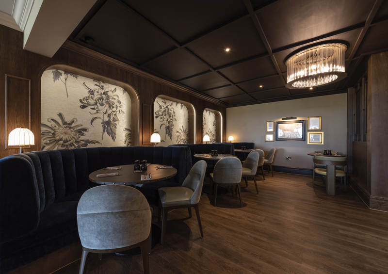 Booth seating at Cook House Bar & Kitchen. Three round dining tables can be seen along the wall to the left of the image. Each has semi-circular banquette seating along the wall, and two chairs on the opposite side, each table is laid with cutlery and napkins. The floor is dark wood, the ceiling is panelled in dark wood and behind each booth is an alcove featuring light wallpaper with a large floral design. There are small lamps between each booth, ceiling spotlights and a large glass light fitting.