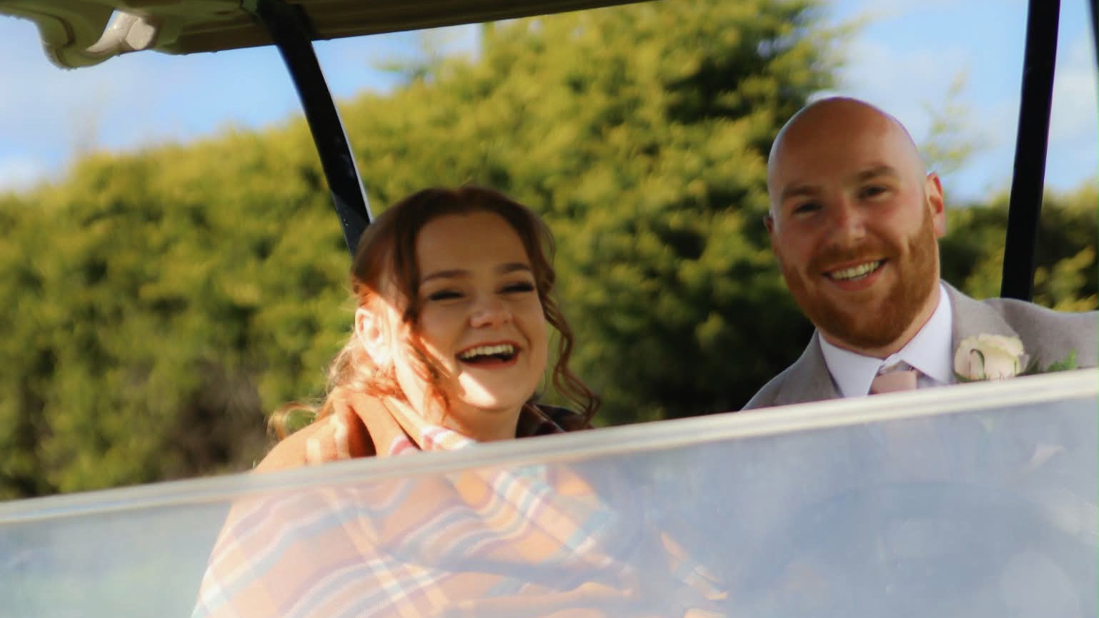 A bride and groom smiling from a green golf buggy featuring The Manor's logo.