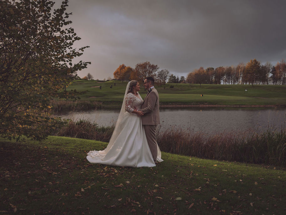 A bride and groom embrace beside The Lake at The Manor Leeds in autumn.