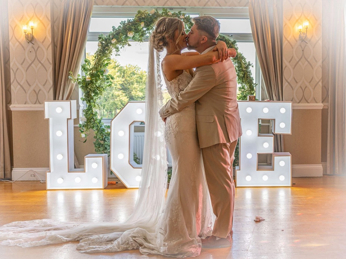 A bride and groom embrace on The Manor's dancefloor in front of a floral hoop and large light-up letters spelling the word "LOVE".
