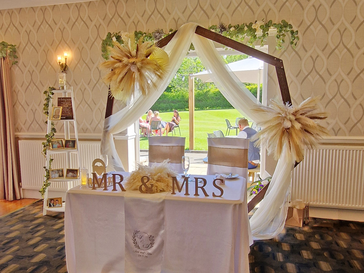 A sweetheart table set for a wedding breakfast in the High Garden room at The Manor. A wooden arch is behind decorated with pampas grass and swagged fabric, and the doors behind are open, with The Lawn and patio seen beyond.