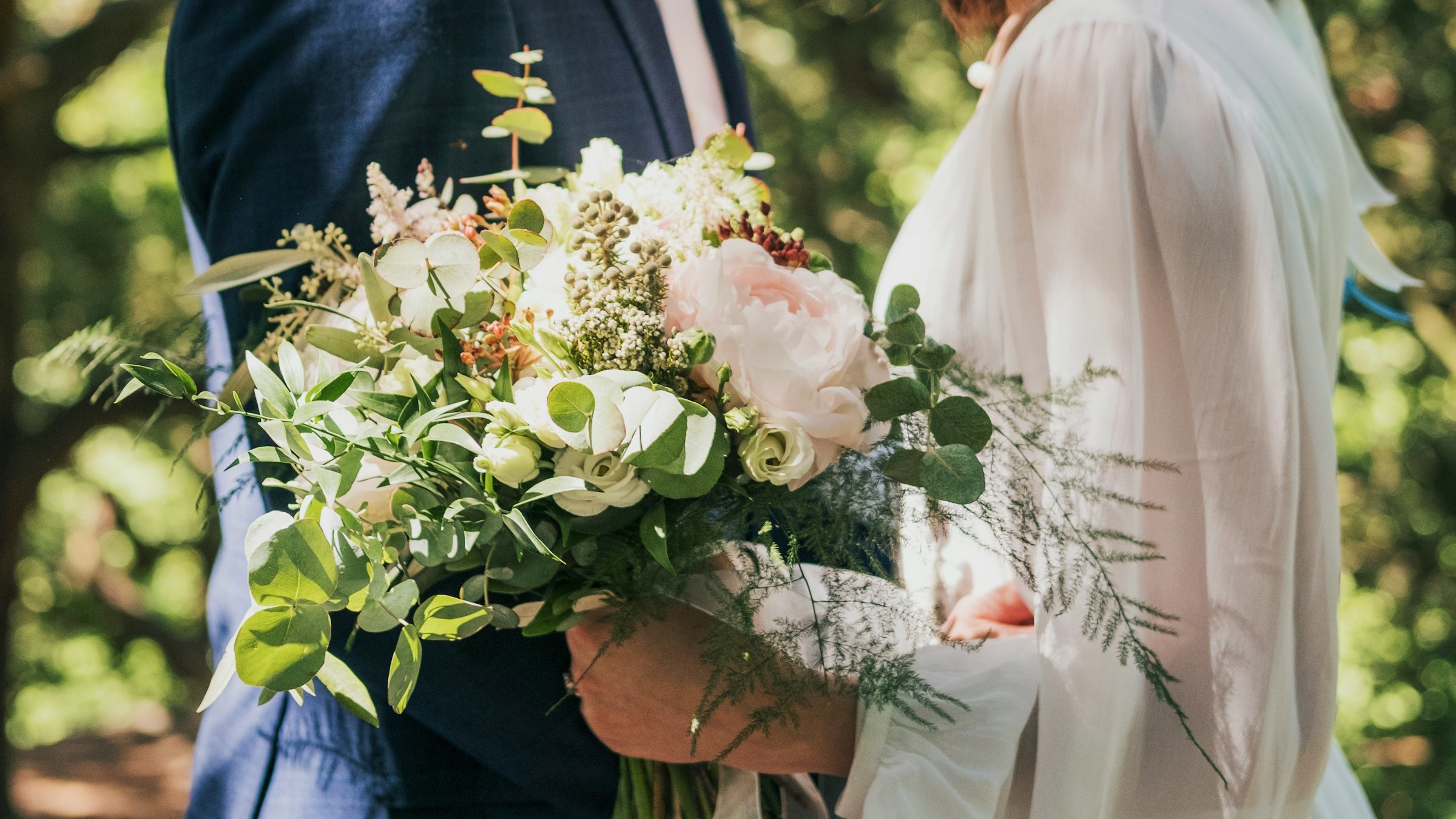 A headless shot of a bride and groom, facing each other. The bride is holding a large, loose bouquet that features pale pink peonies, white roses and eucalyptus.