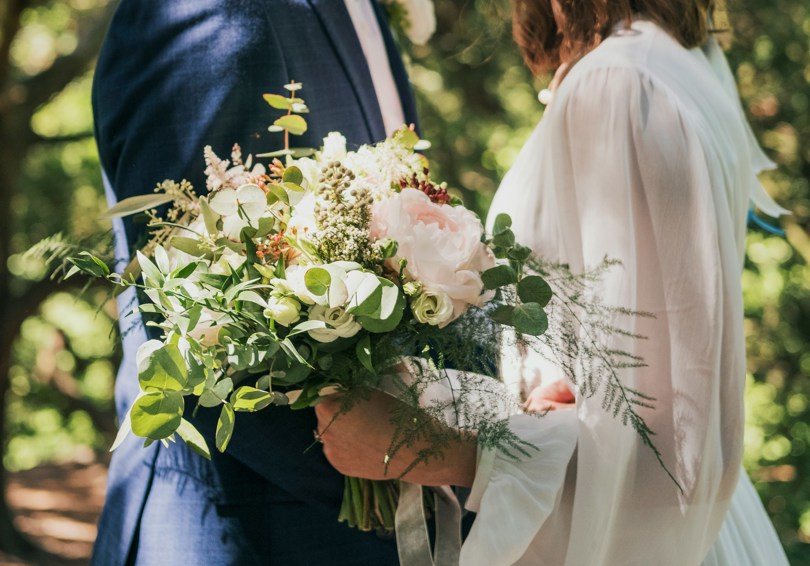 A headless shot of a bride and groom, facing each other. The bride is holding a large, loose bouquet that features pale pink peonies, white roses and eucalyptus.