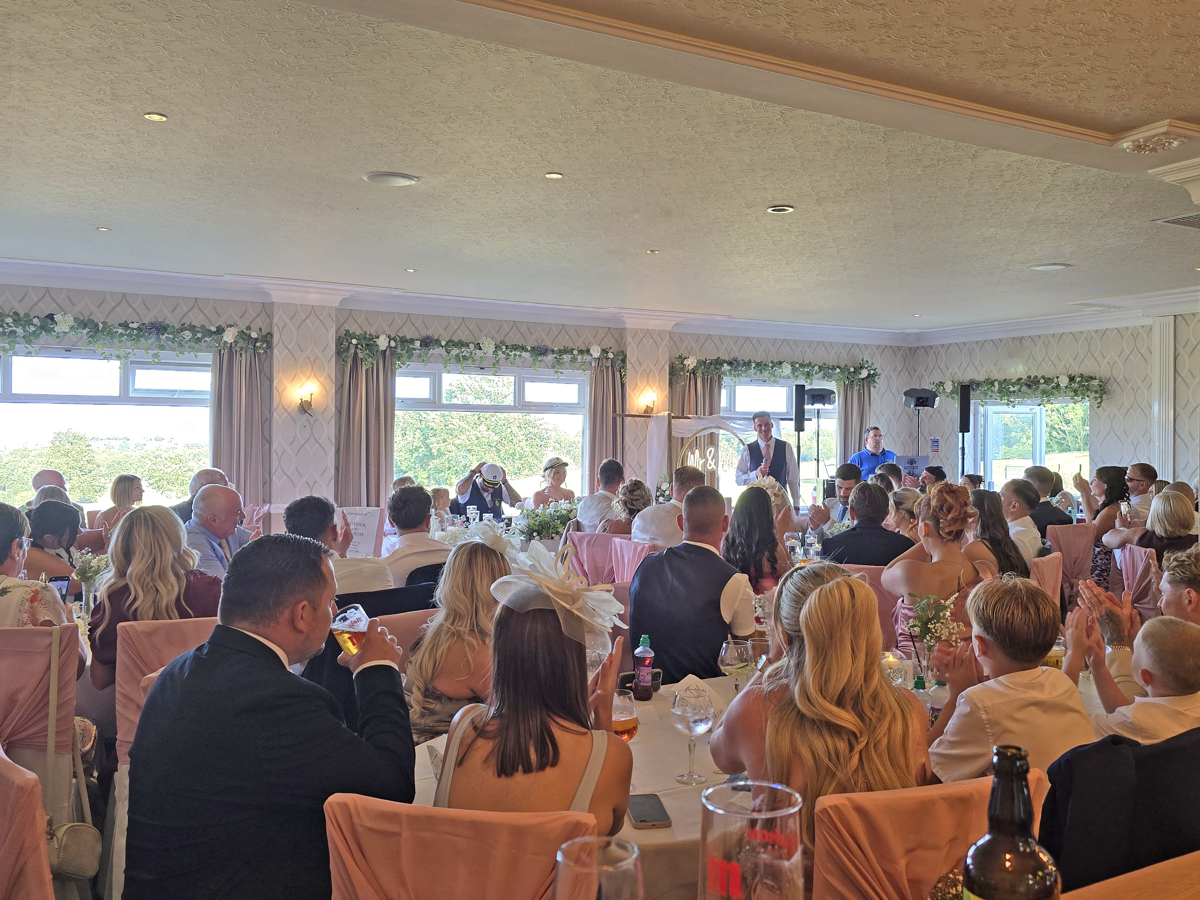 Wedding guests are seated at tables in The View as a Best Man makes a speech. The bride and groom are sitting a sweetheart table on the dancefloor facing their guests.