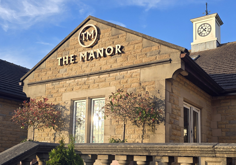 The main entrance of The Manor, showing the stone balustrade, the clocktower and the gold logo.