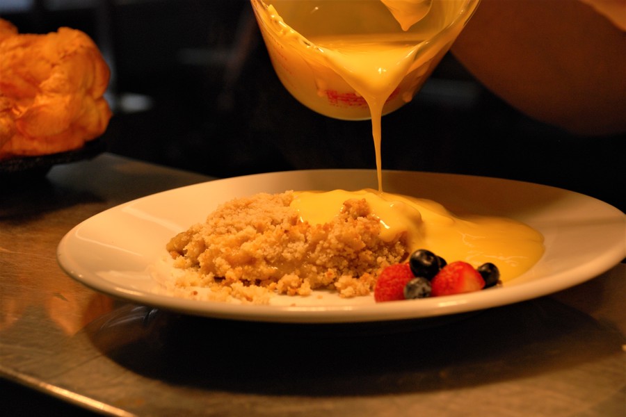 Custard being poured over crumble on a plate with berries on the side.