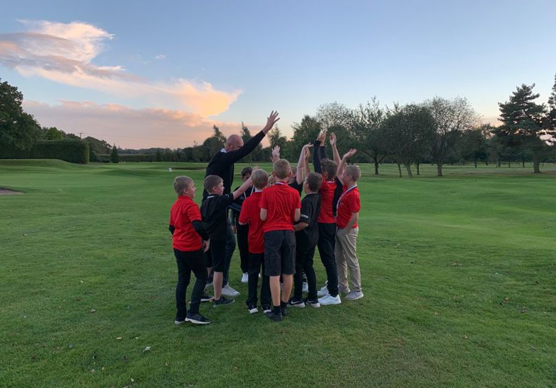 A group of young golfers with golf coach Mark McGeehan, most wear red shirts and have their arms up in celebration.