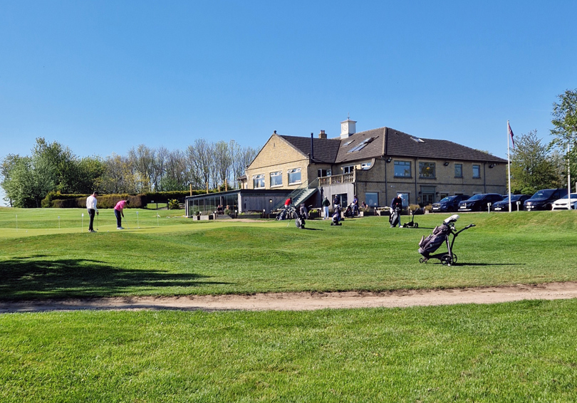A view of the putting green at The Manor Leeds, looking towards the main building and Cook House patio and terrace. There are a number of golfers practicing.