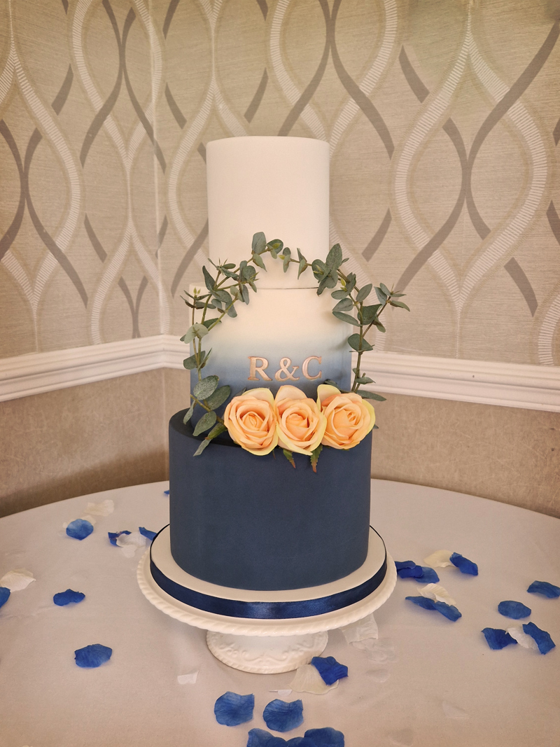 A three tier wedding cake on a table in High Garden at The Manor. The bottom tier is dark blue, the top tier is white and the middle tier is a gradient blending the two, three peach roses and "R & C" decorate the middle.