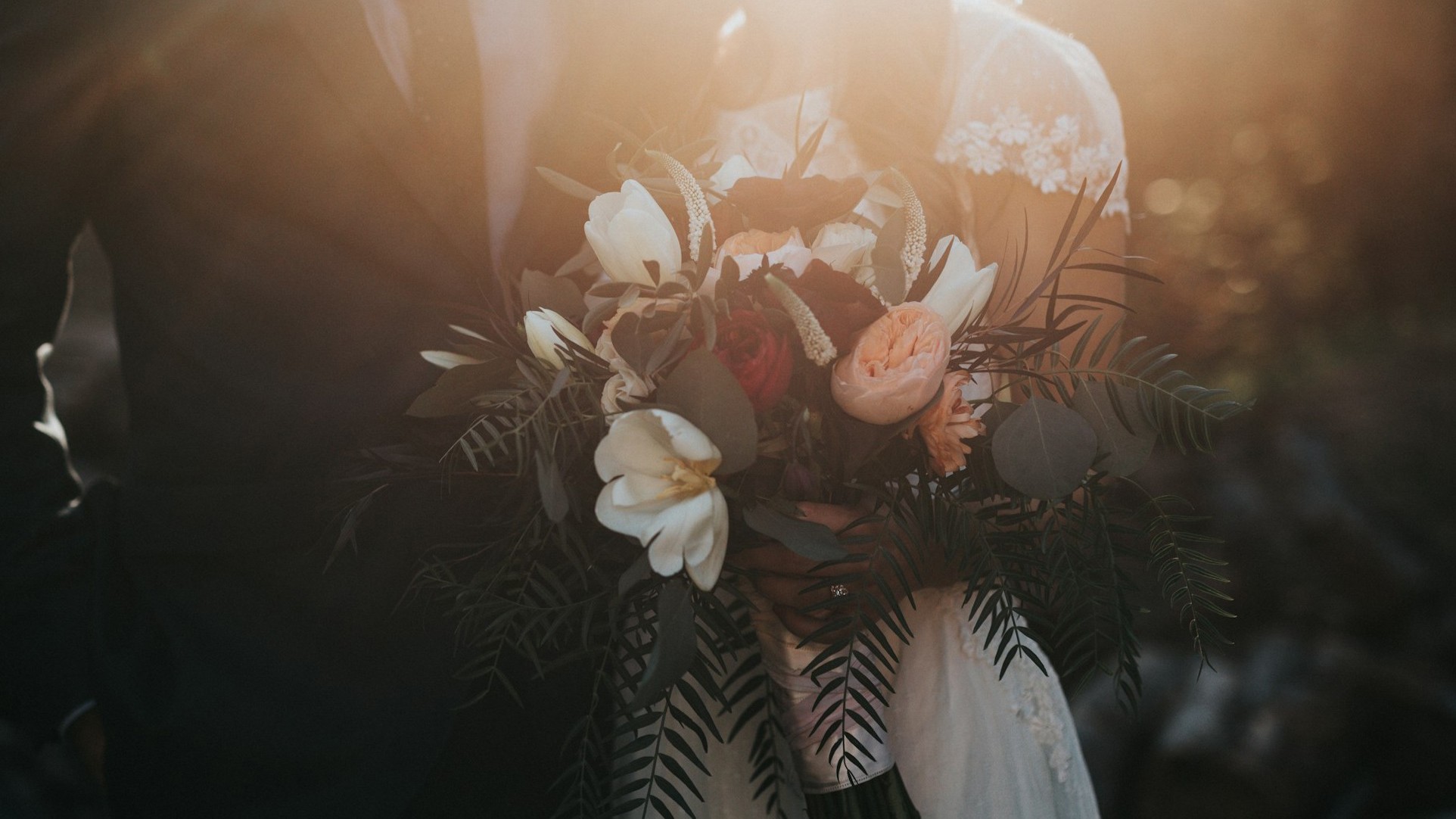 A headless shot of a bride and groom, taken at sunset. The bride is holding a bouquet that features peach-coloured peonies and palm fronds.