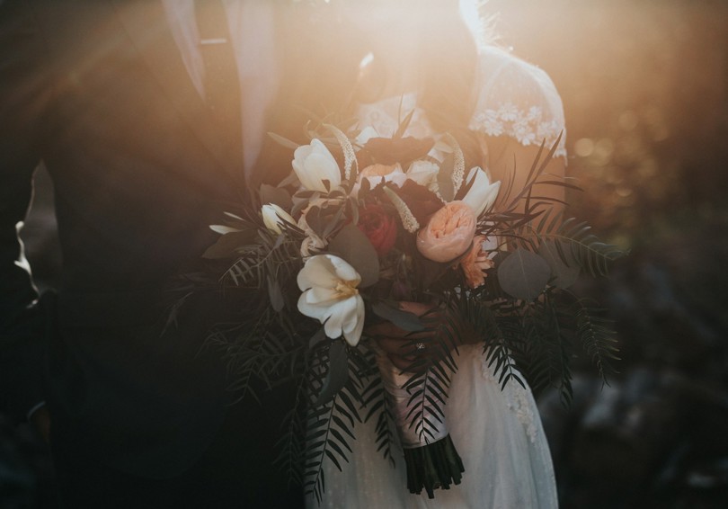 A headless shot of a bride and groom, taken at sunset. The bride is holding a bouquet that features peach-coloured peonies and palm fronds.