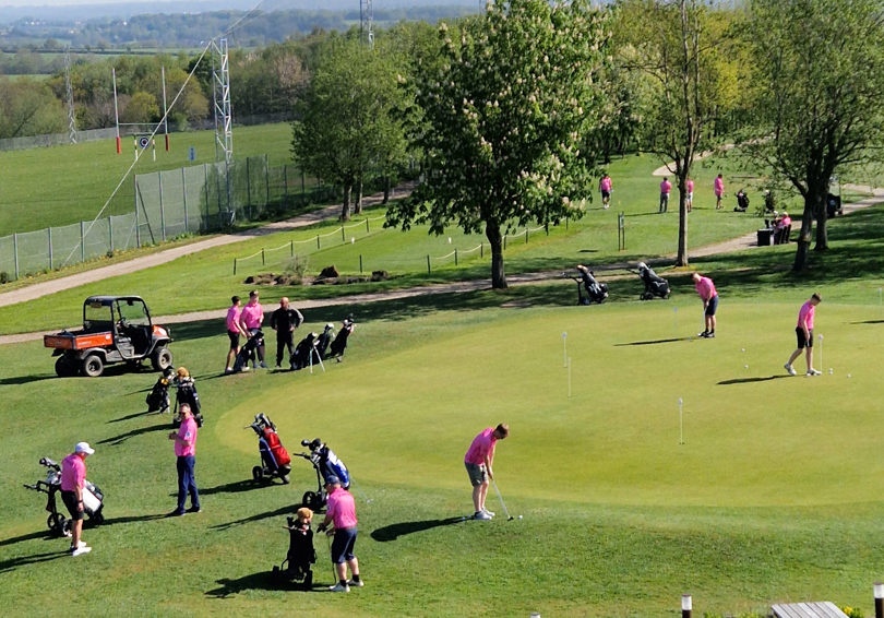 A high view of the putting green, first tee and driving range taken from The Manor's main building. There are a number of golfers, all wearing pink shirts.